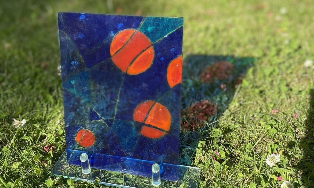 eine blau, rot bemalte Glasplatte steht auf dem Rasen und wirft einen bunten Schatten