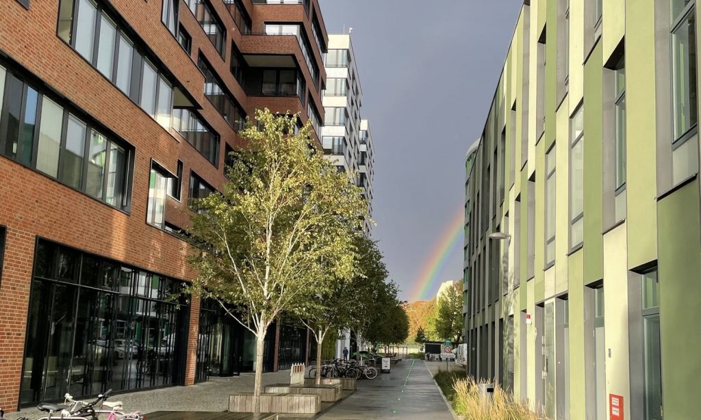 Zwei Gebäude auf dem EUREF-Campus in Berlin, zwischen denen eine kleine Straße verläuft. Am Ende dieser Straße ist ein Regenbogen.
